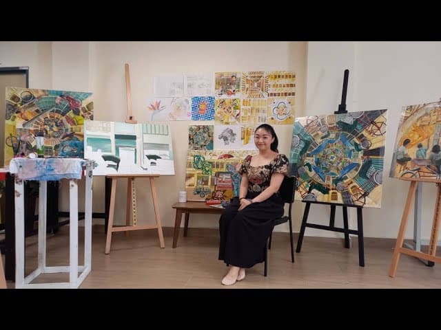 A woman in a paint-splattered apron holds a brush in a bright art studio filled with canvases and supplies
