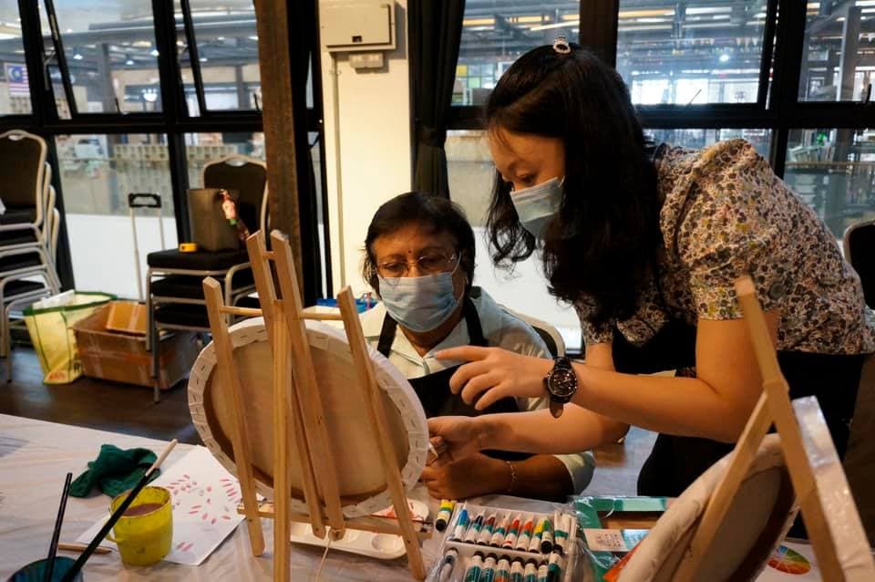 Two women wearing face masks collaborate on a canvas painting at an easel indoors.