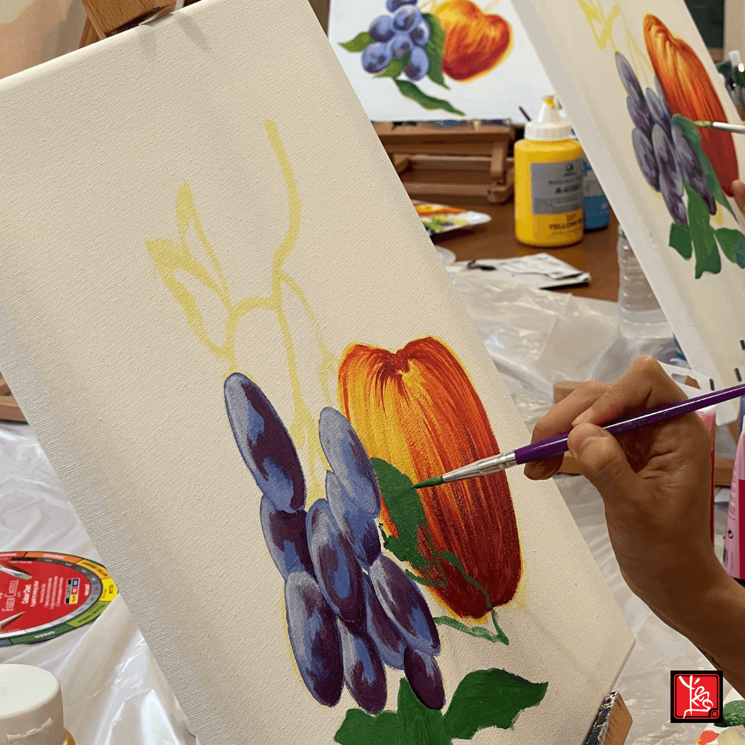 Hand painting purple grapes and a red apple on a white canvas with a paintbrush.