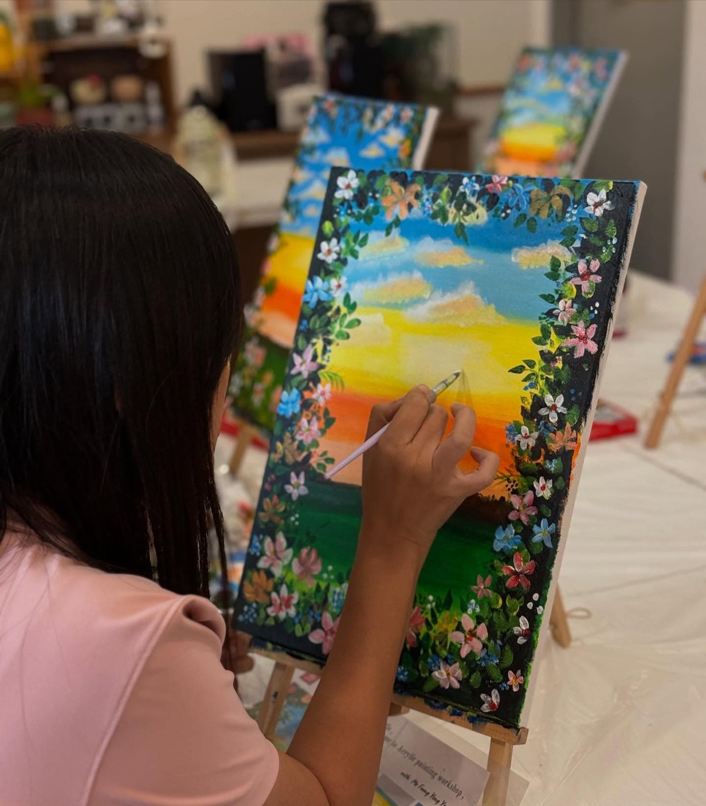 Person painting a vibrant sunset landscape with a floral border on a canvas in a workshop.
