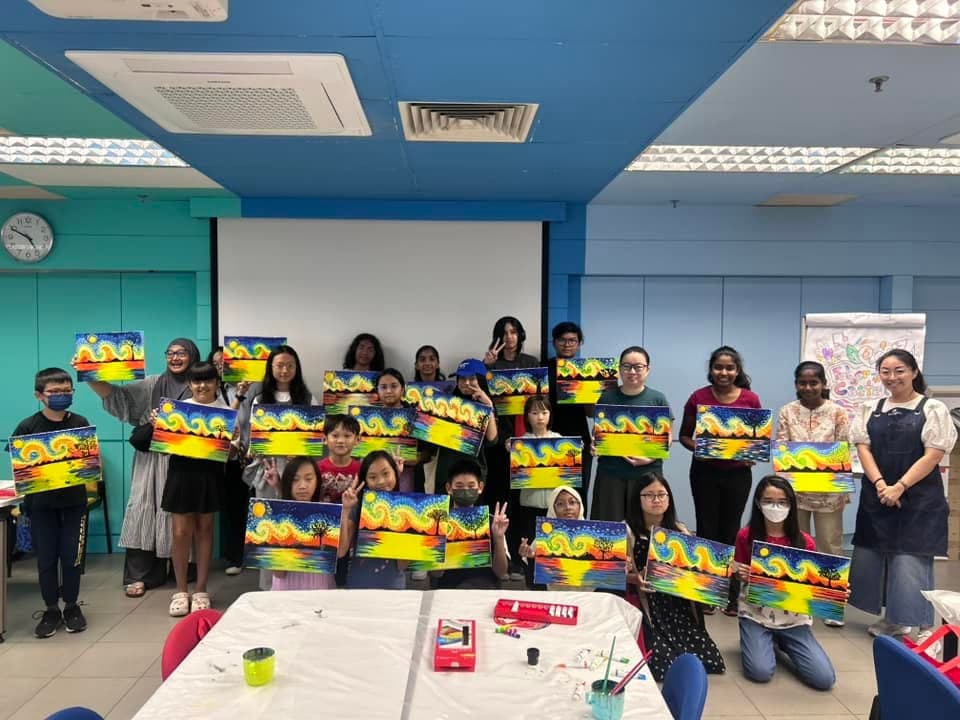 Diverse students and teachers holding their vibrant sunset landscape paintings in a blue classroom.