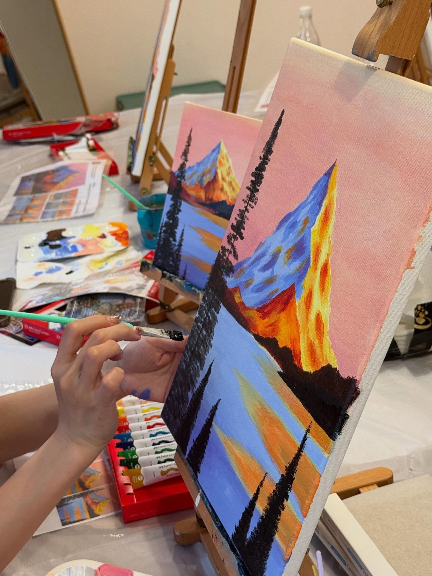 A person painting a vibrant mountain landscape with black pine trees on a canvas easel.