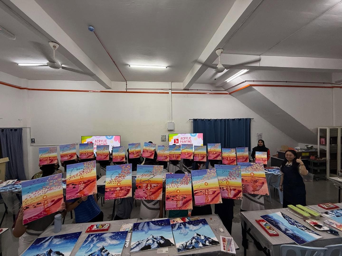 Participants at an acrylic painting workshop display their finished sunset and mountain landscape canvases.