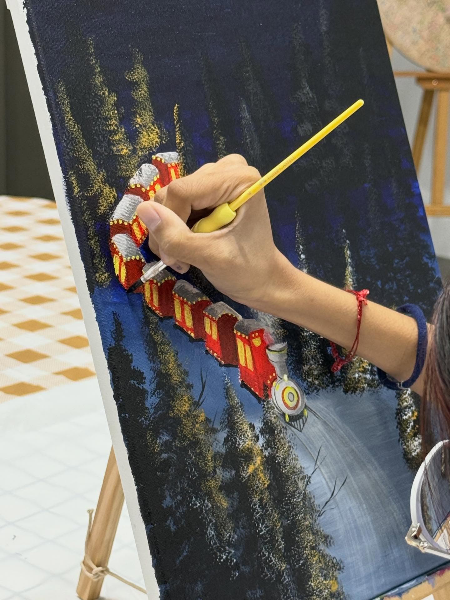 Hand painting a red train traveling through a dark, snowy forest on a canvas easel.