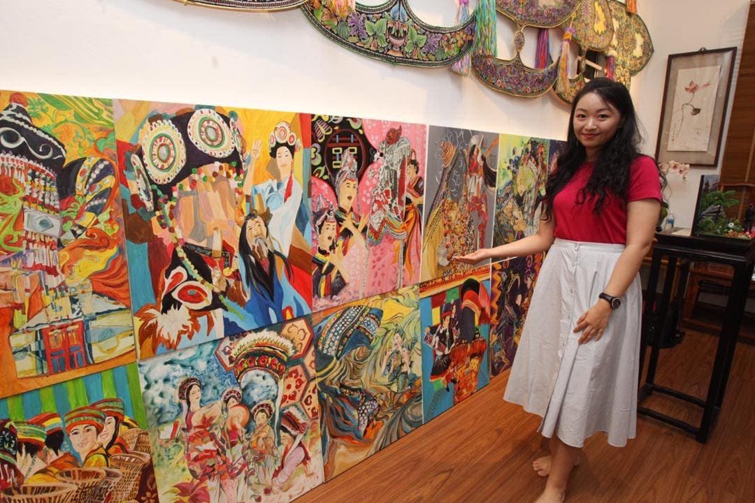 Woman in red shirt standing next to a wall of colorful, traditional Chinese-style paintings.