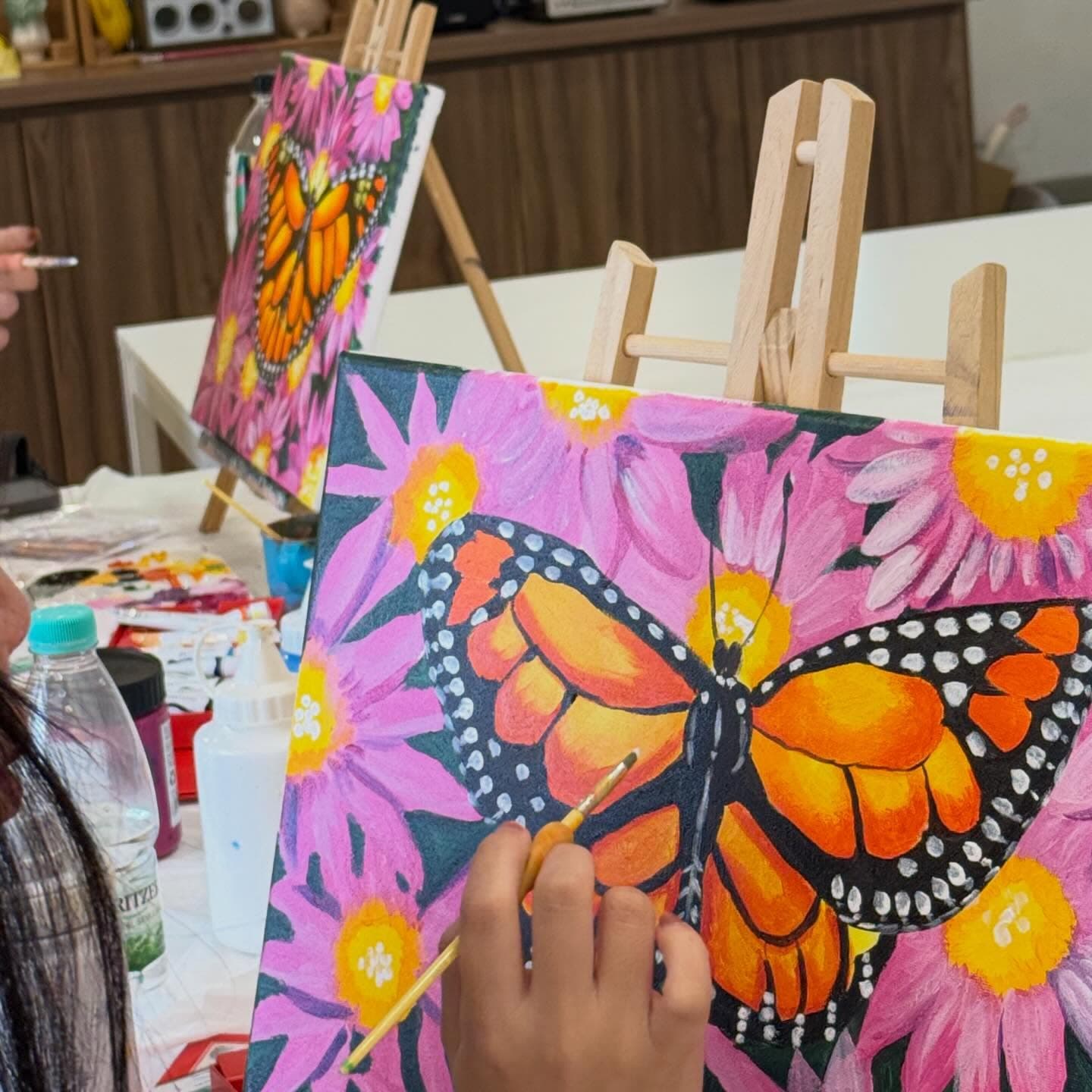 Hand painting a monarch butterfly on a canvas with pink flowers during an art class.