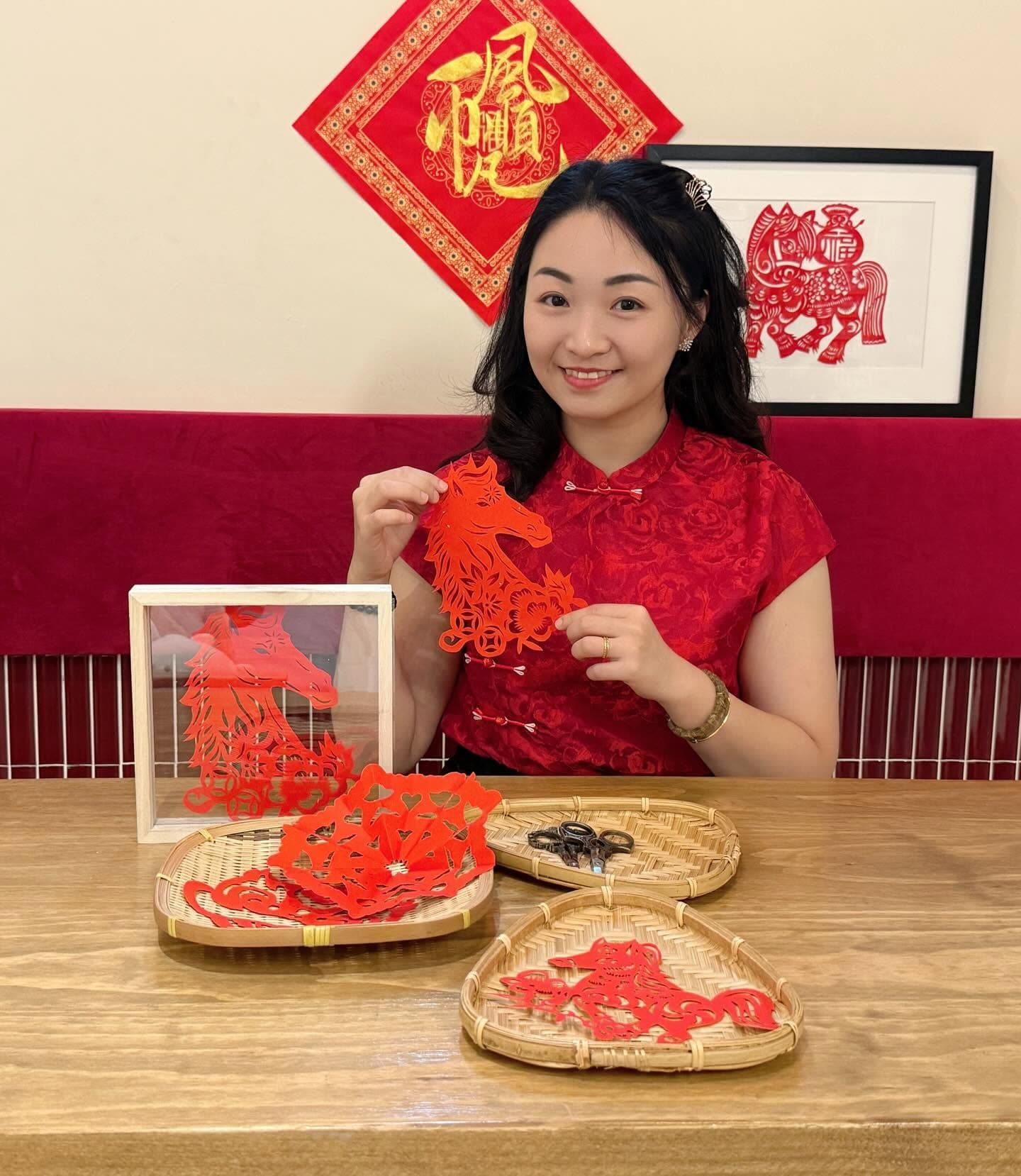 Smiling woman in red traditional dress holding a red paper-cut horse art piece.