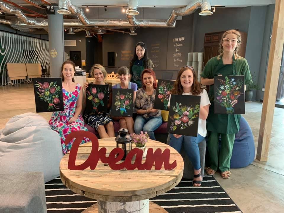 Seven women smiling and holding their floral paintings in a modern industrial art studio.
