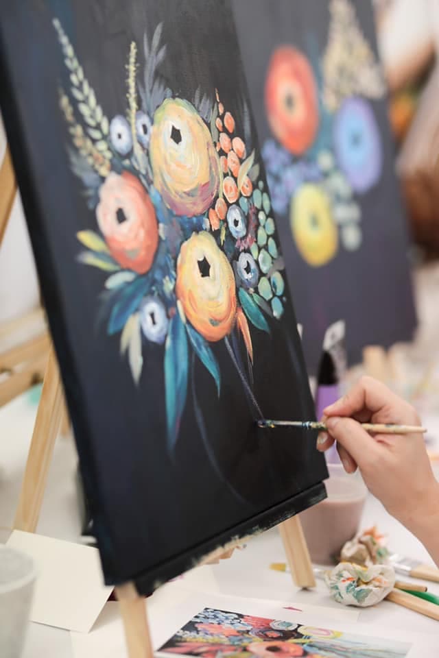 A hand paints vibrant, impressionistic flowers on a black canvas with a fine-tipped brush.