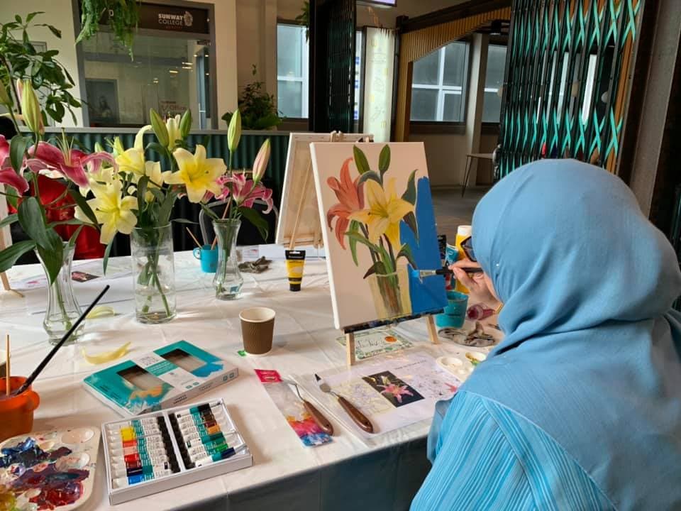 Artist in a blue hijab paints lilies on a canvas using real flowers as a reference.