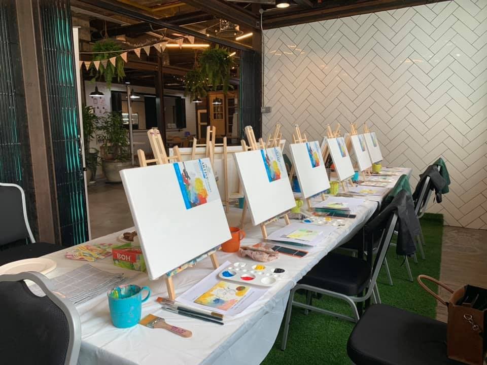 Row of blank canvases on easels with paint palettes and brushes on a long table.