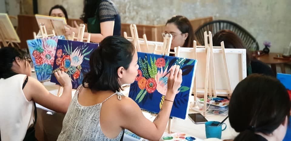 Women painting colorful floral designs on canvases at easels during a group art workshop.