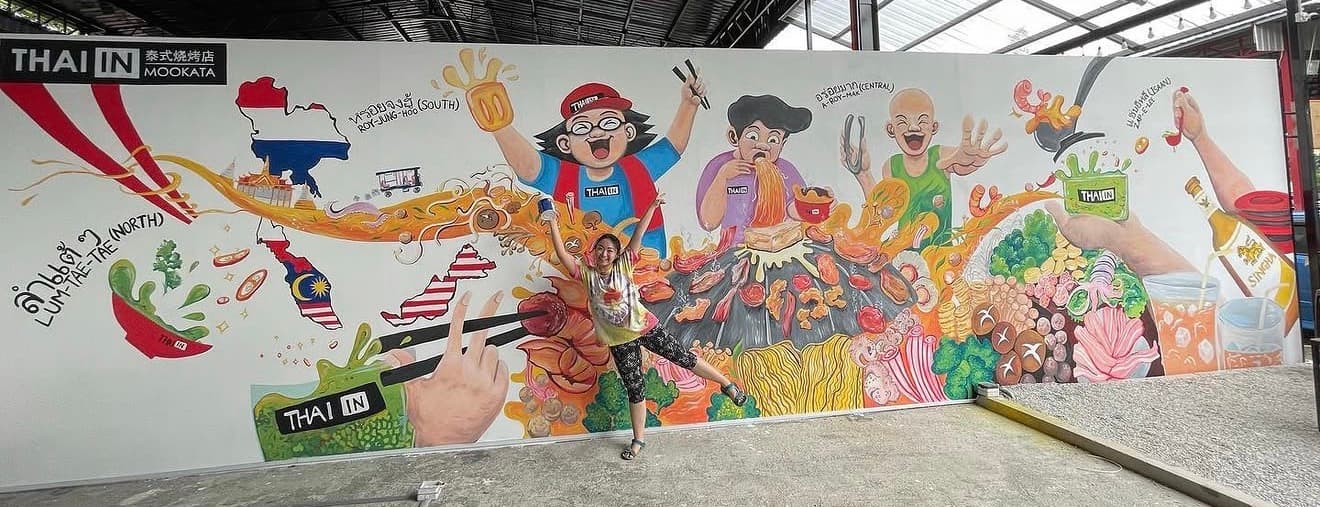 Woman posing in front of a vibrant mural featuring cartoon characters eating Thai mookata barbecue.