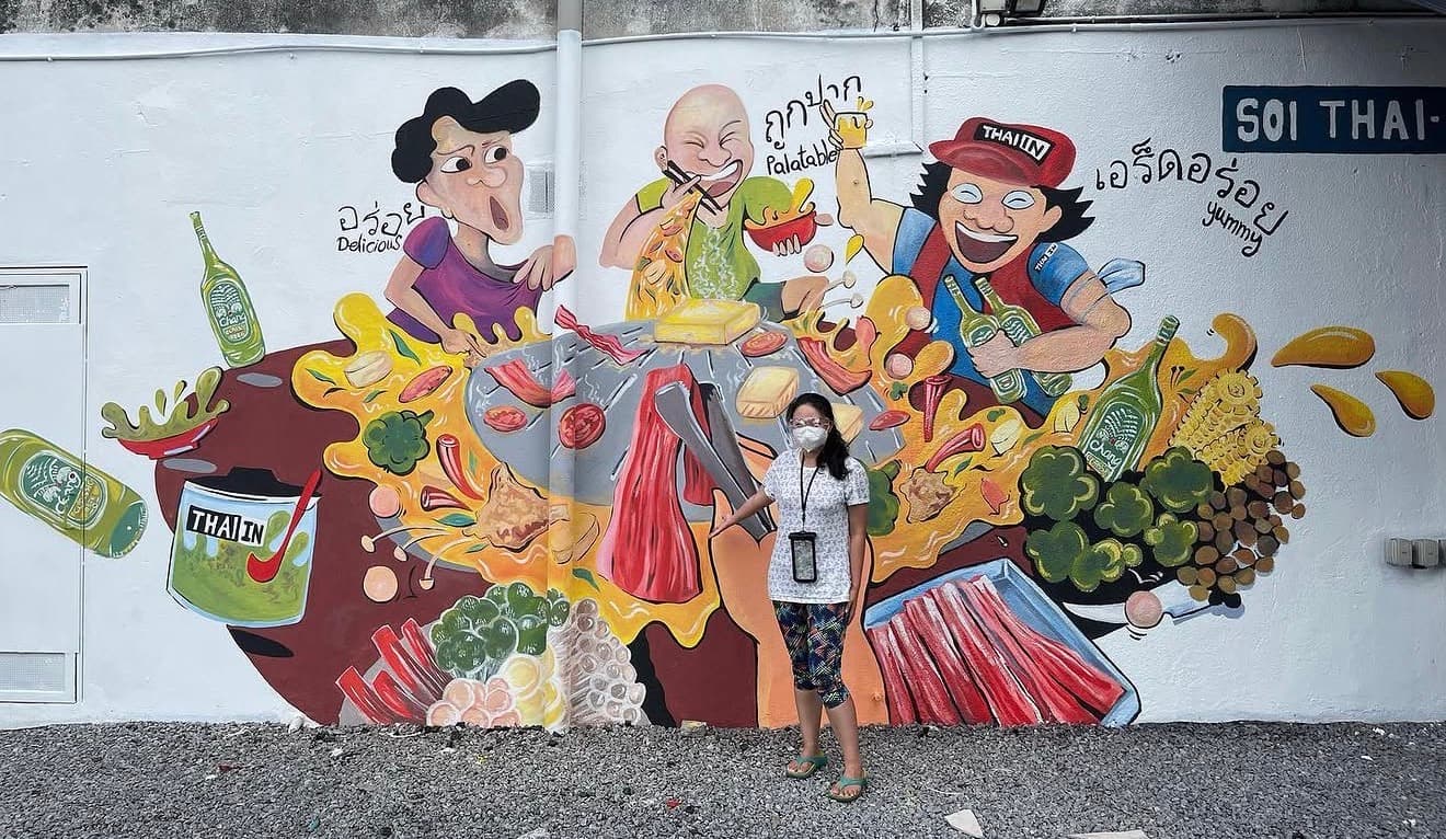 Woman stands in front of a vibrant mural depicting cartoon characters eating Thai barbecue.