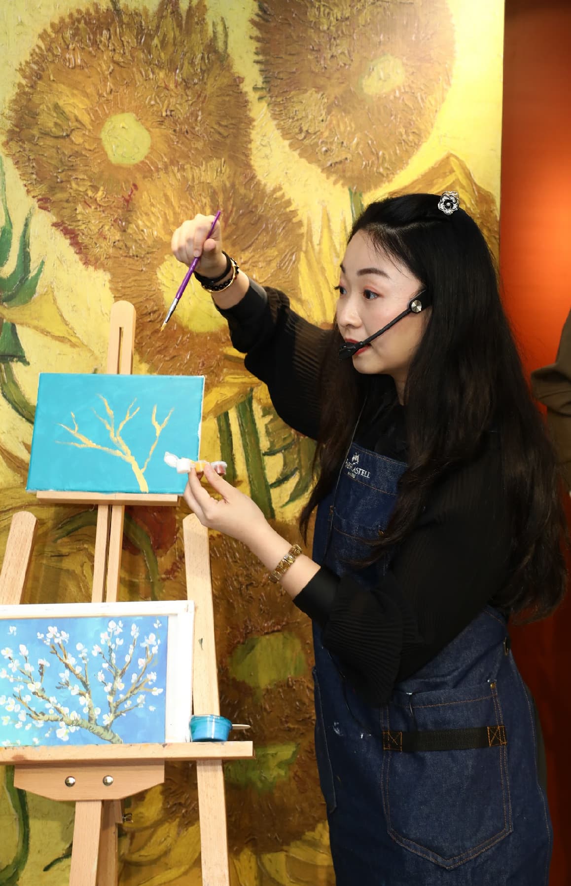 Woman in a denim apron paints on a canvas against a large sunflower painting backdrop.