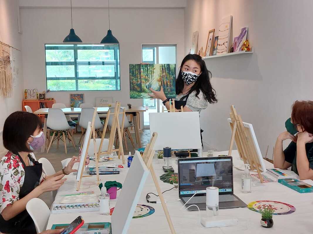 Masked art instructor shows a forest painting to students at easels in a studio.
