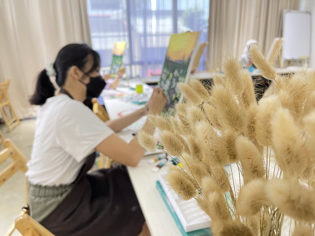 Masked woman paints a landscape on canvas behind fuzzy dried grass in the foreground.