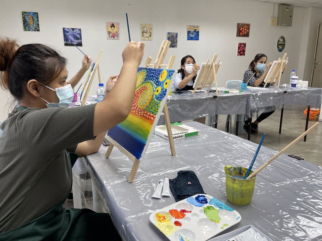 Masked students painting colorful canvases on wooden easels in a bright art studio classroom.