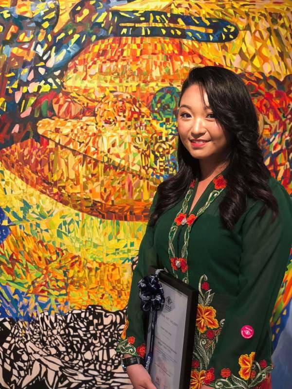 Smiling woman in green embroidered kebaya holding a framed award before a vibrant, colorful painting.